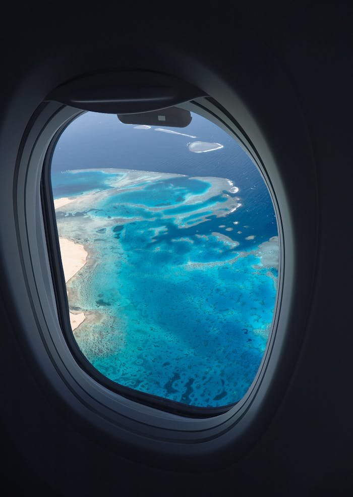 Breathtaking aerial view of ocean reefs through an airplane window, showcasing vibrant blues from above.